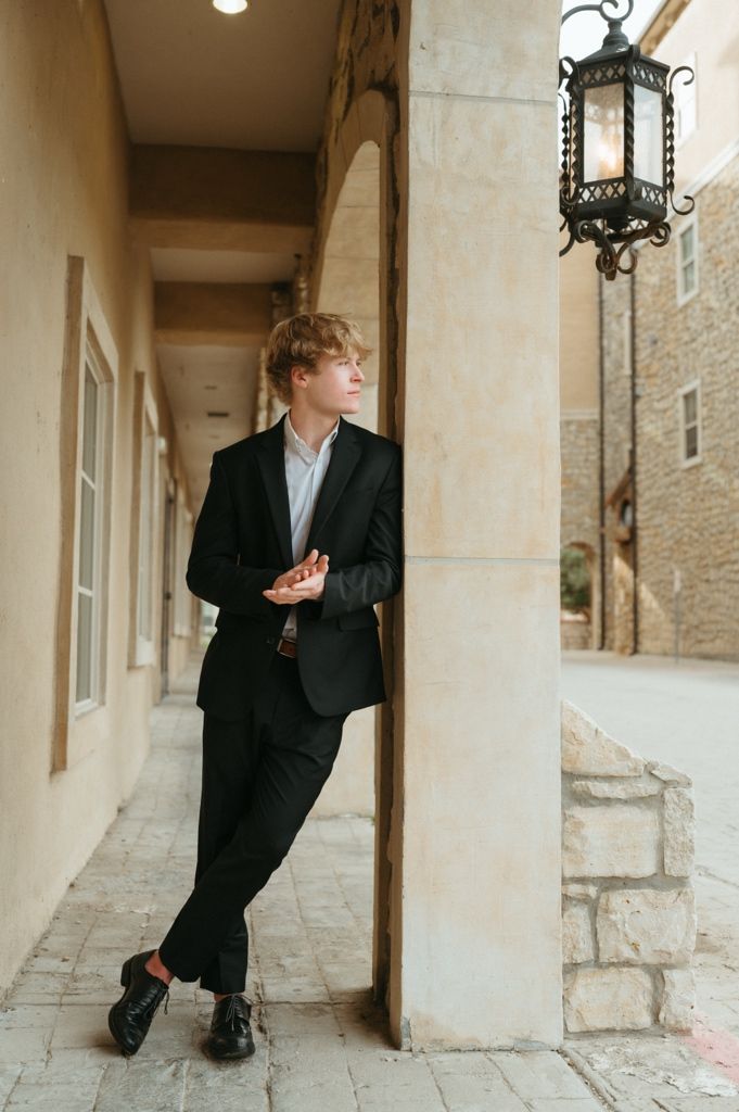 Young man in black suit leaning on pillar in archway, vintage lantern overhead, full body shot.