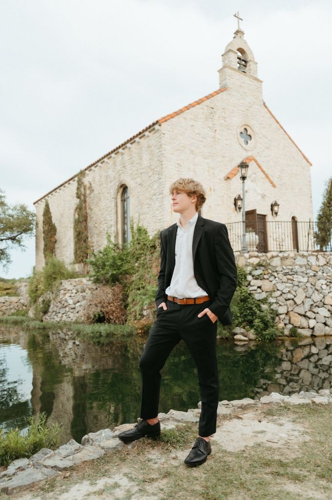 Young man in black suit standing by reflective pond with historic stone chapel in background.