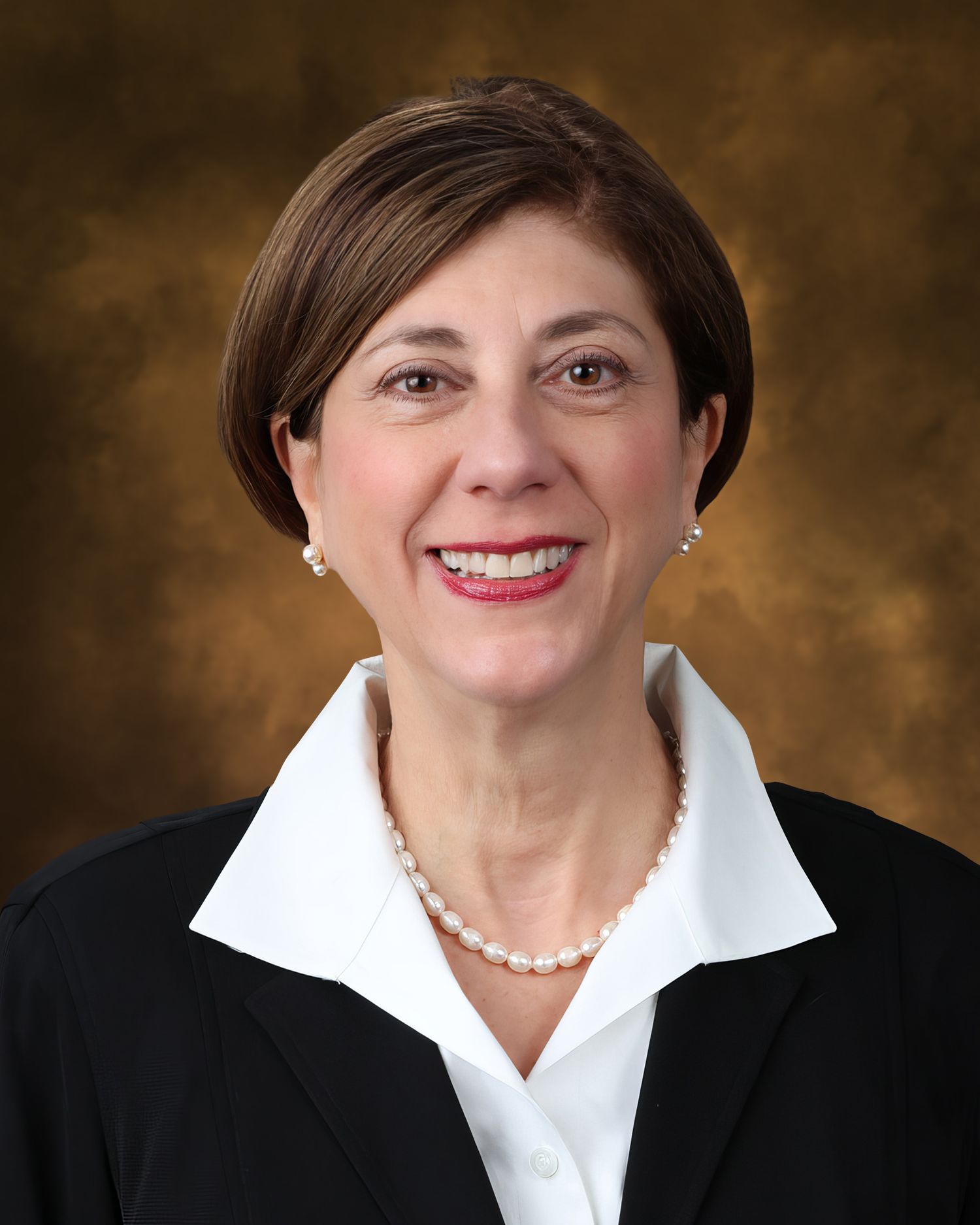 Professional headshot of a smiling woman in a black blazer, white blouse, and pearl necklace against a brown background.