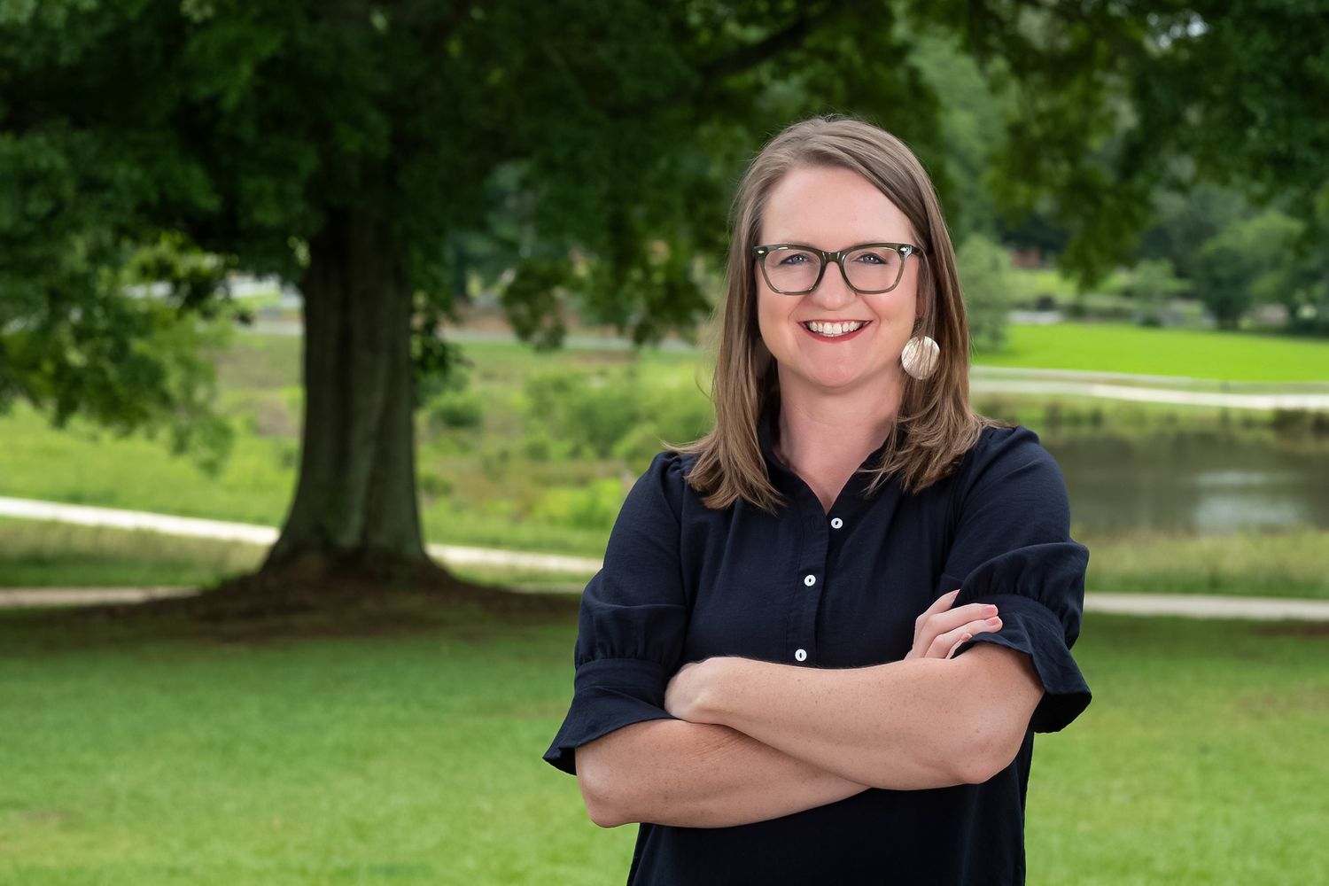 Smiling woman with glasses stands arms crossed outdoors near a large tree in a lush green park setting.