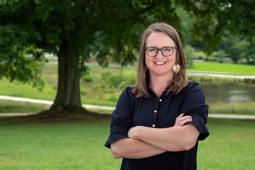 Smiling woman with glasses stands arms crossed outdoors near a large tree in a lush green park setting.