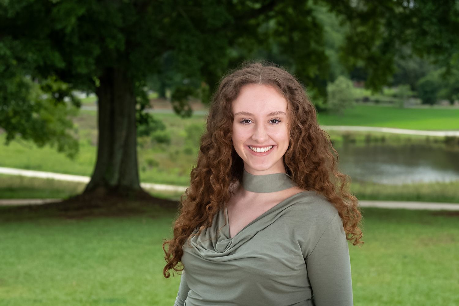 Smiling young woman with curly hair in a green top posing outdoors in a lush park setting.