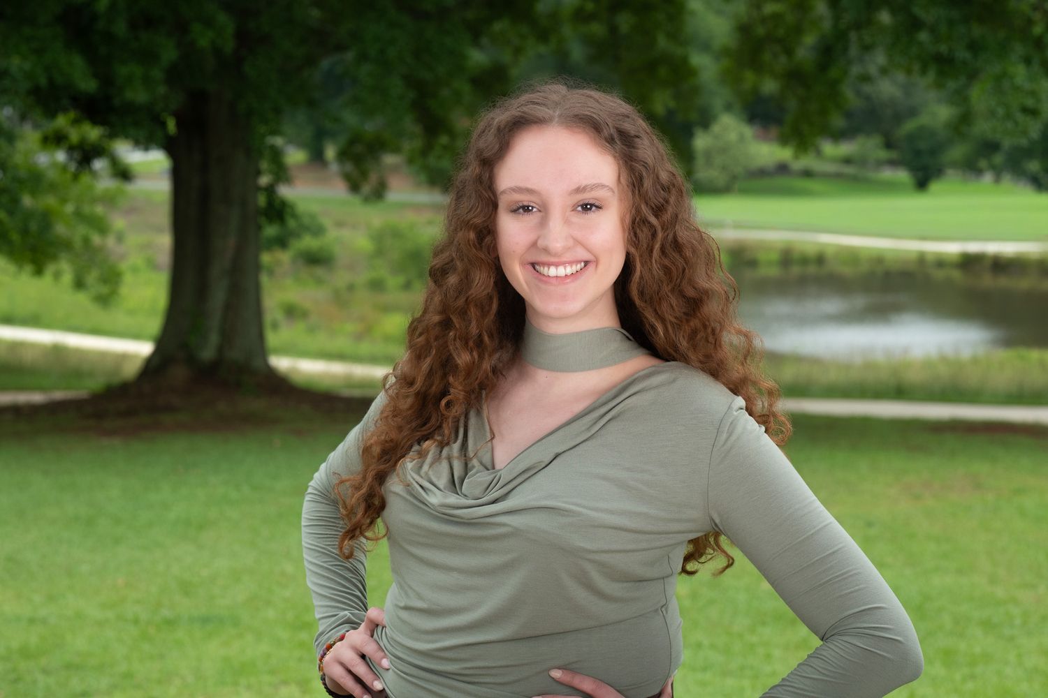 Professional Headshot Session with a young woman with curly hair smiling outdoors in a green top with hands on hips near a scenic pond.