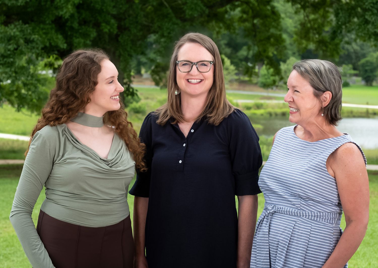 Three women smiling outdoors in a lush green park setting, dressed in casual summer attire.