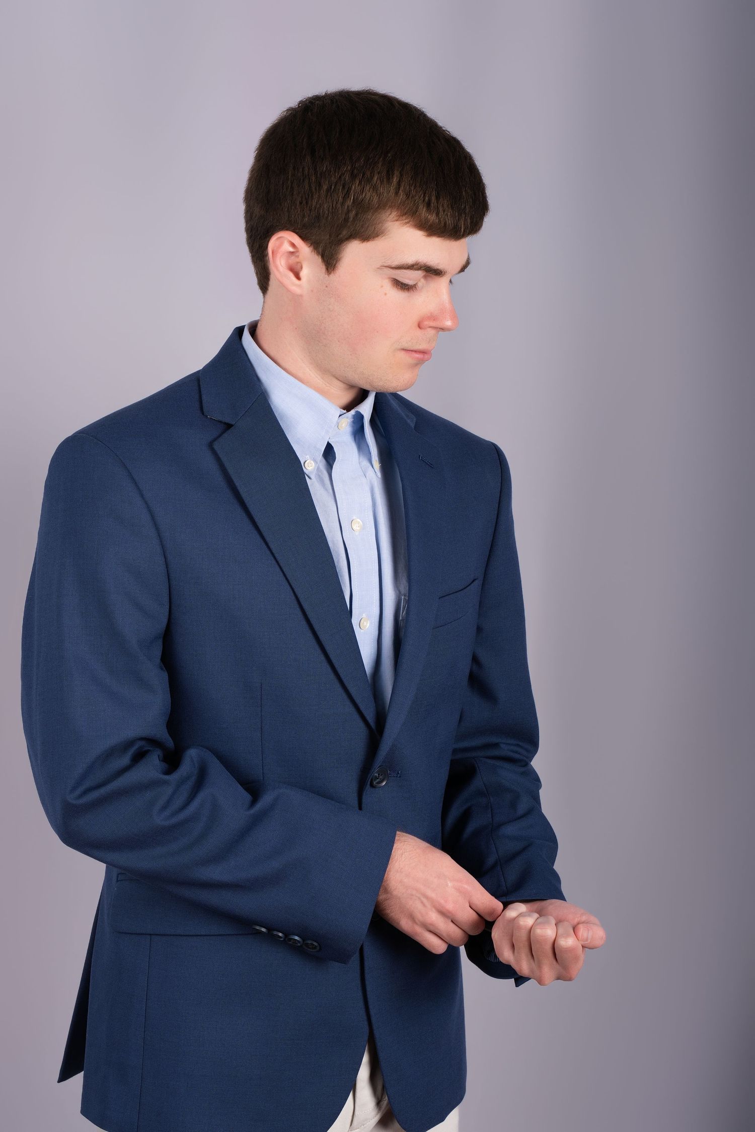 Professional Headshot Session with a young man in a navy suit adjusting his sleeve while looking down against a gray studio background.