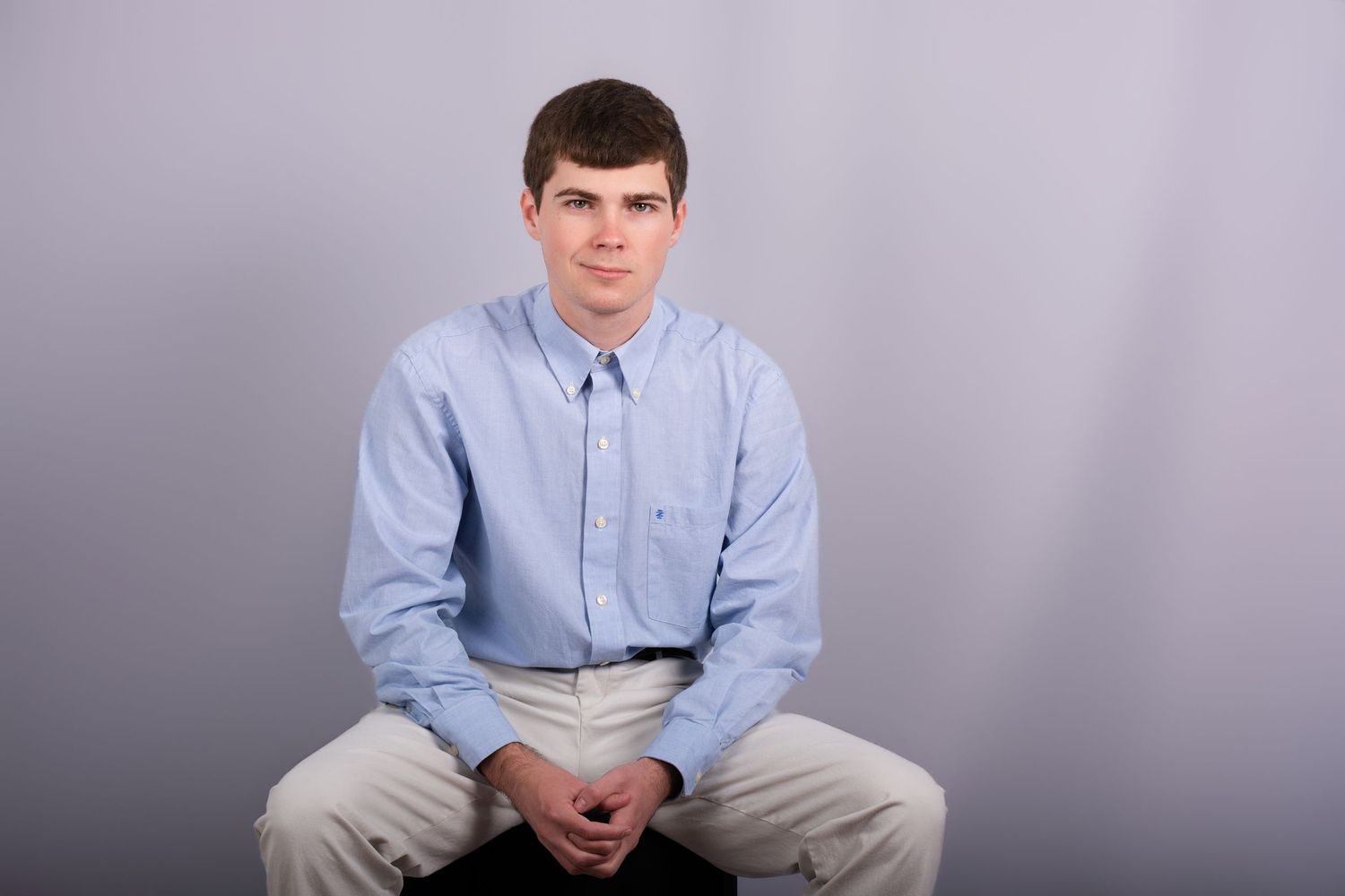 Professional Headshot Session with a young man in a light blue button-down shirt seated and posing against a plain gray background.