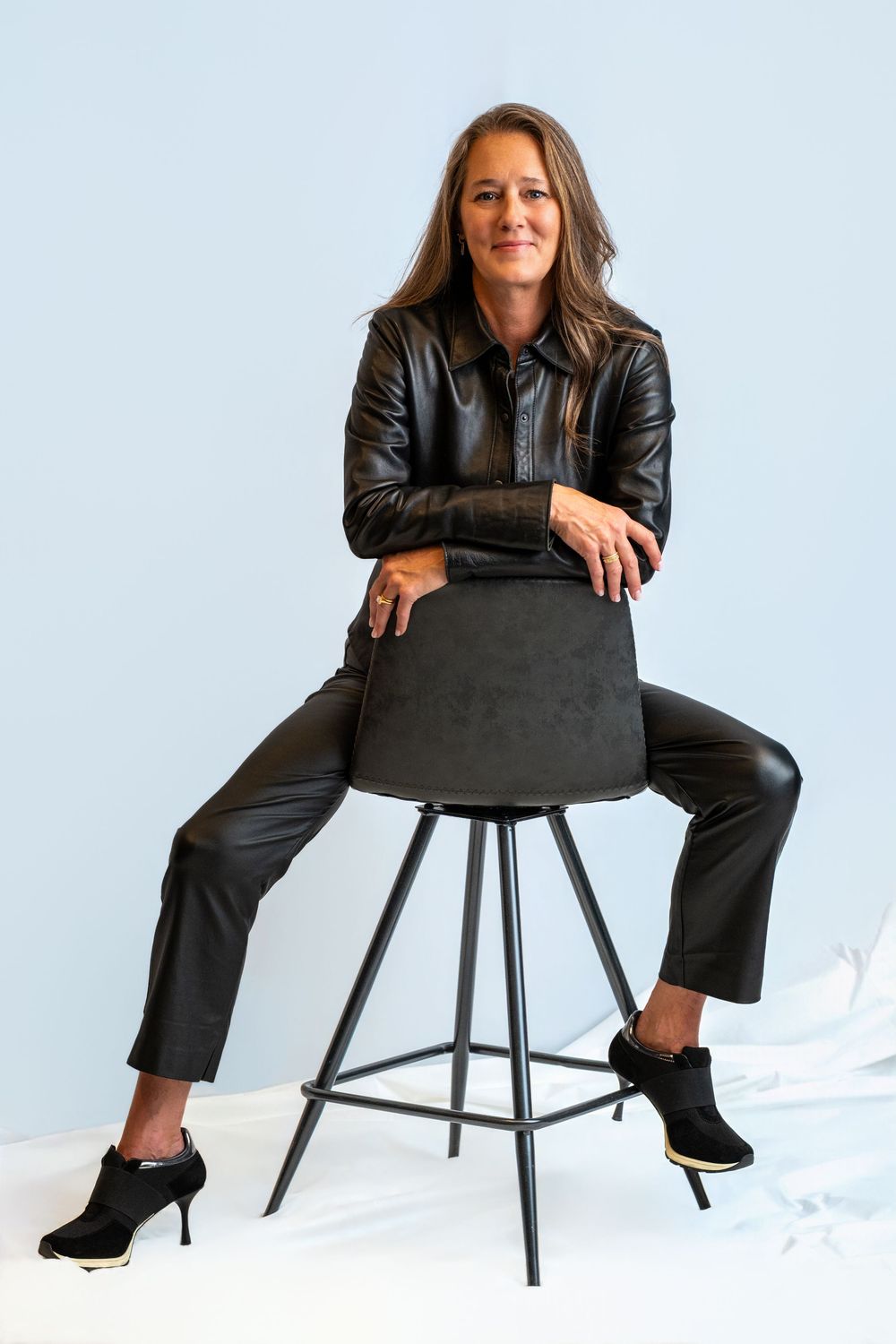 Woman in a black leather jacket and pants sits astride a black barstool, leaning on its back, against a light blue studio backdrop.