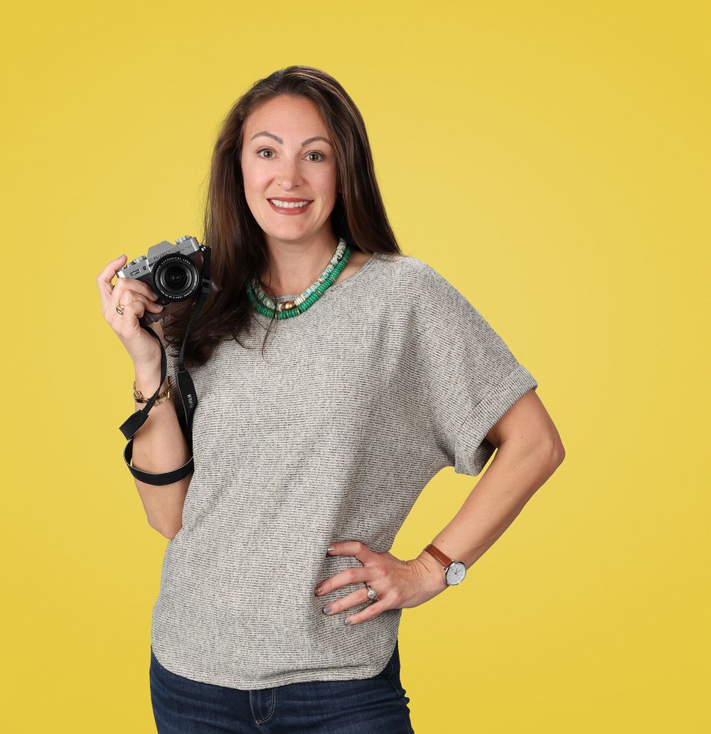 Smiling woman holding a camera with a green necklace and gray top poses against a yellow background.