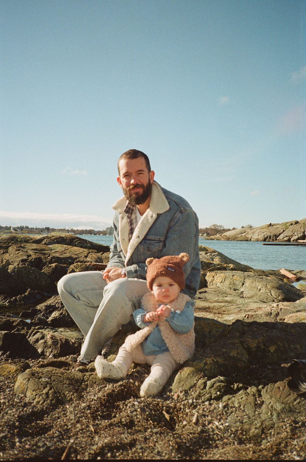 Bearded father sitting on rocky beach with his baby wearing a cute bear hat on sunny day.