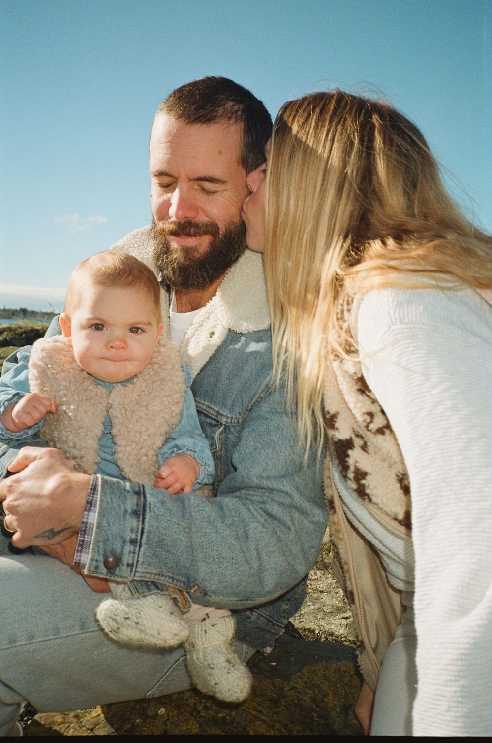 Happy couple sharing a tender kiss moment while holding their cute baby on a sunny beach.