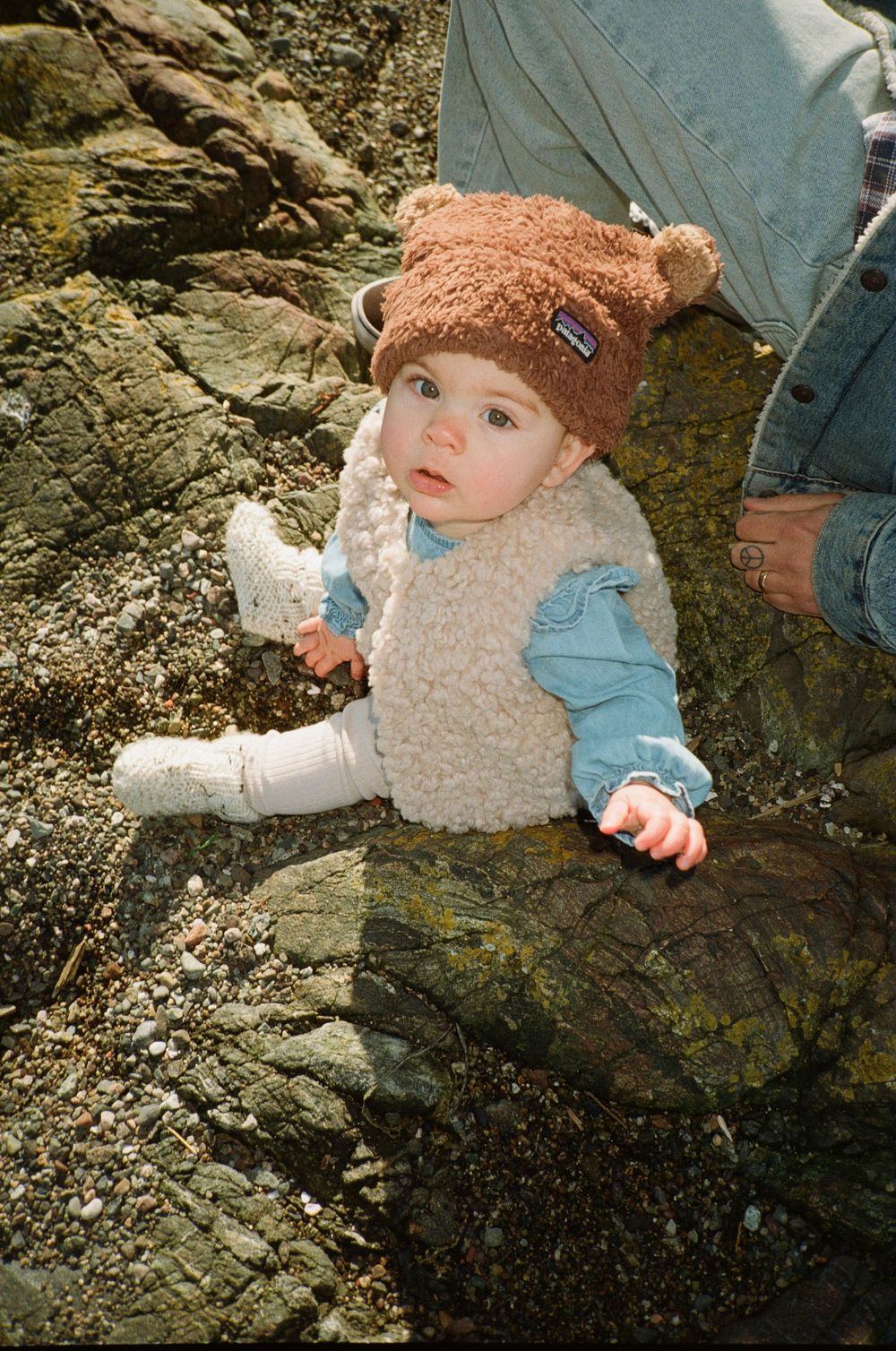 Adorable baby in bear hat and fluffy vest sitting on coastal rocks on a sunny day.