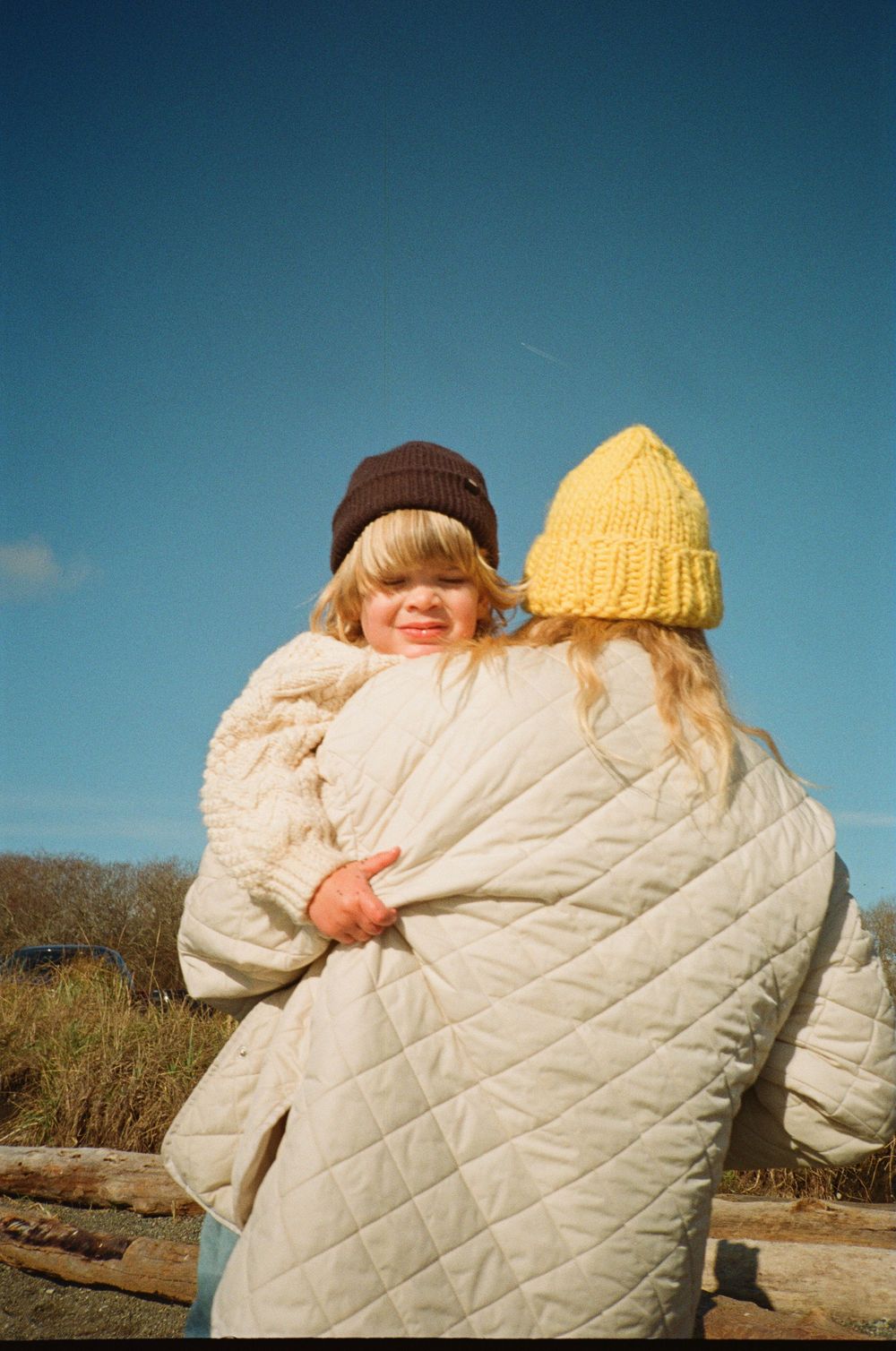 Two children hugging outdoors wrapped in a white quilted blanket wearing colorful winter beanies.