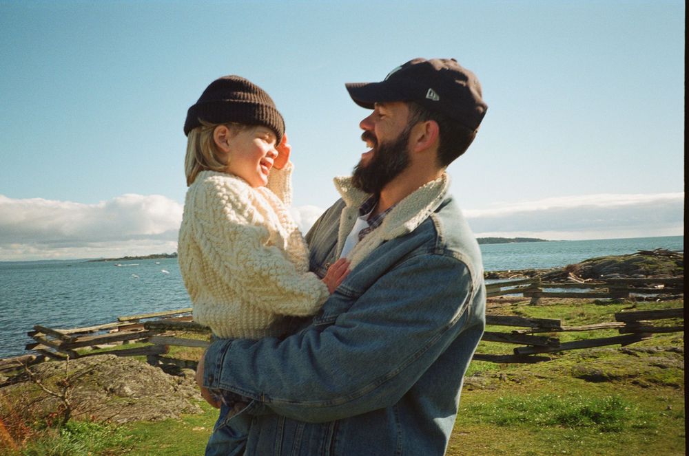 Bearded man in denim jacket lifting smiling child near ocean on a bright sunny day.