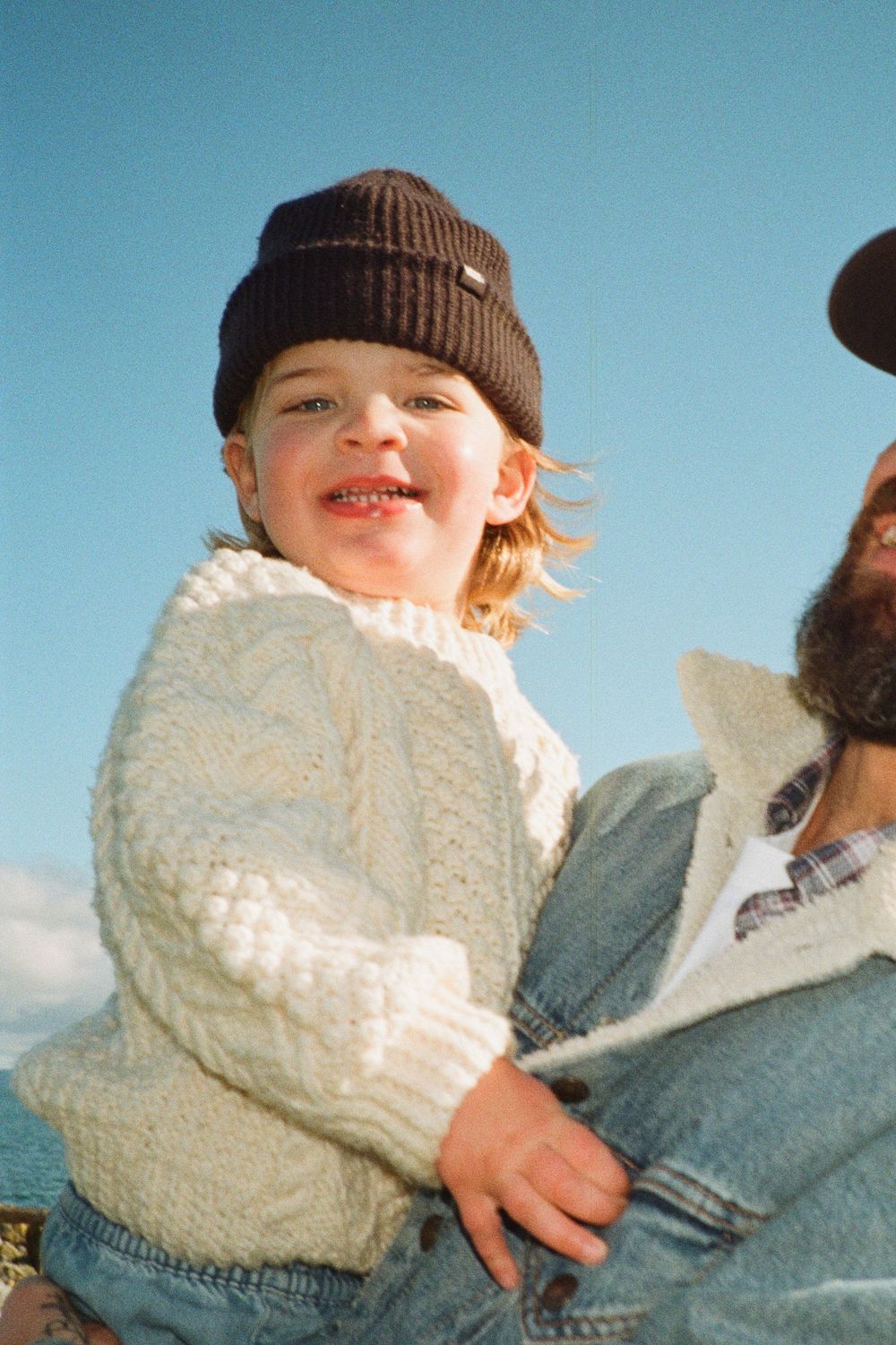 Smiling toddler in dark knit beanie and cream cable-knit sweater held by bearded man outdoors.