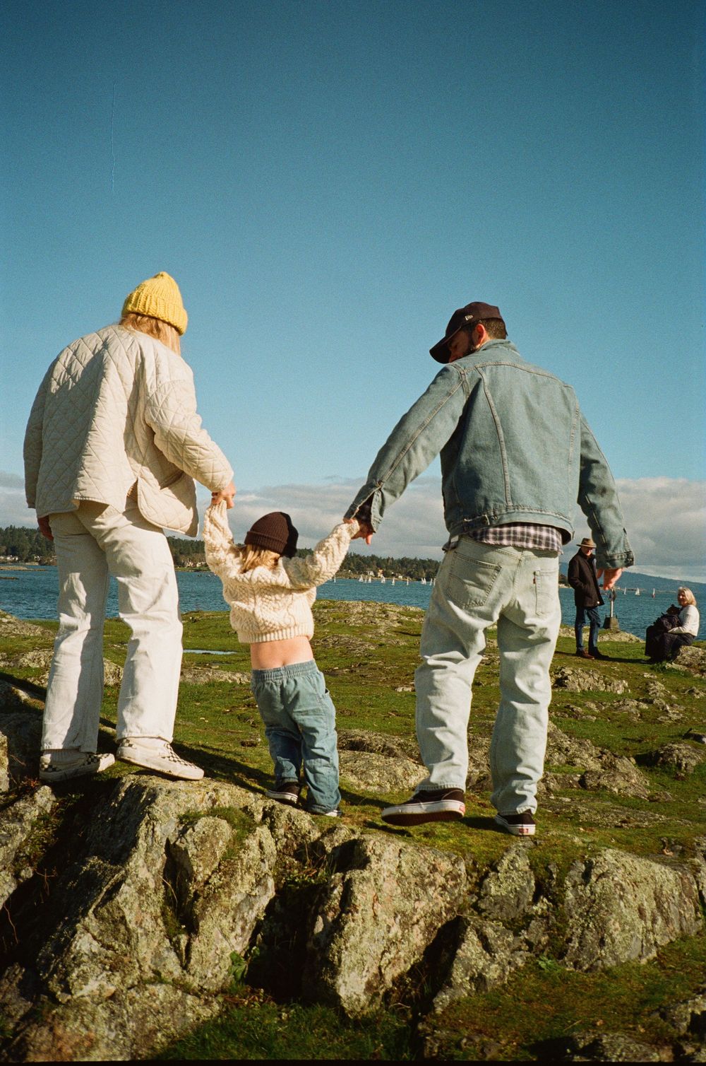 Family of three holding hands on rocky coastal terrain, viewed from behind on sunny day.