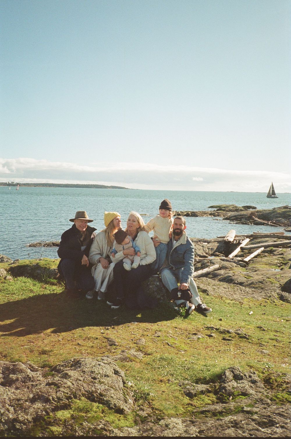 Group of six family members posing together on rocky shore with sailboat in background.