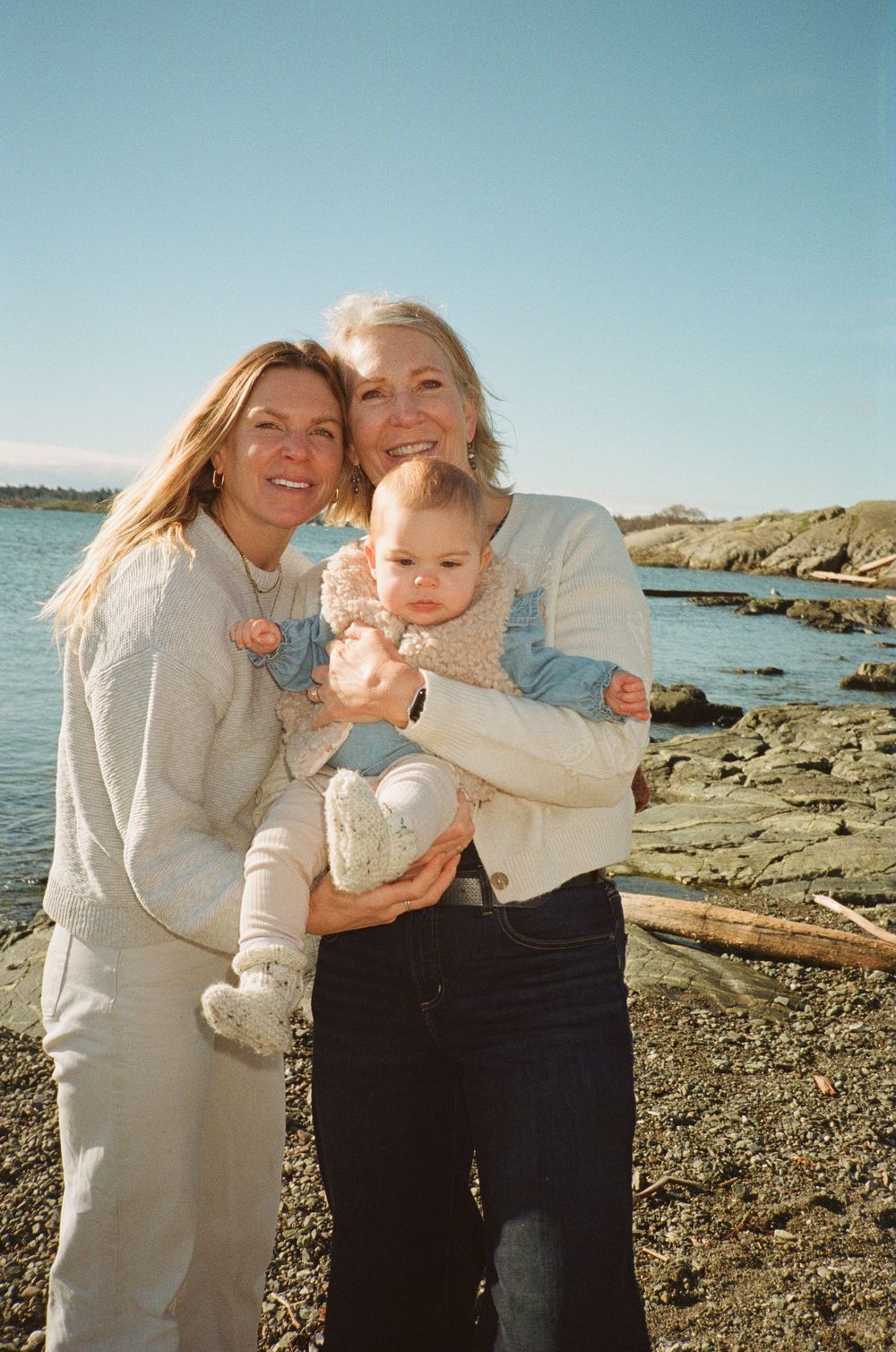 Two women smiling and holding an adorable baby on a sunny rocky beach coastline.