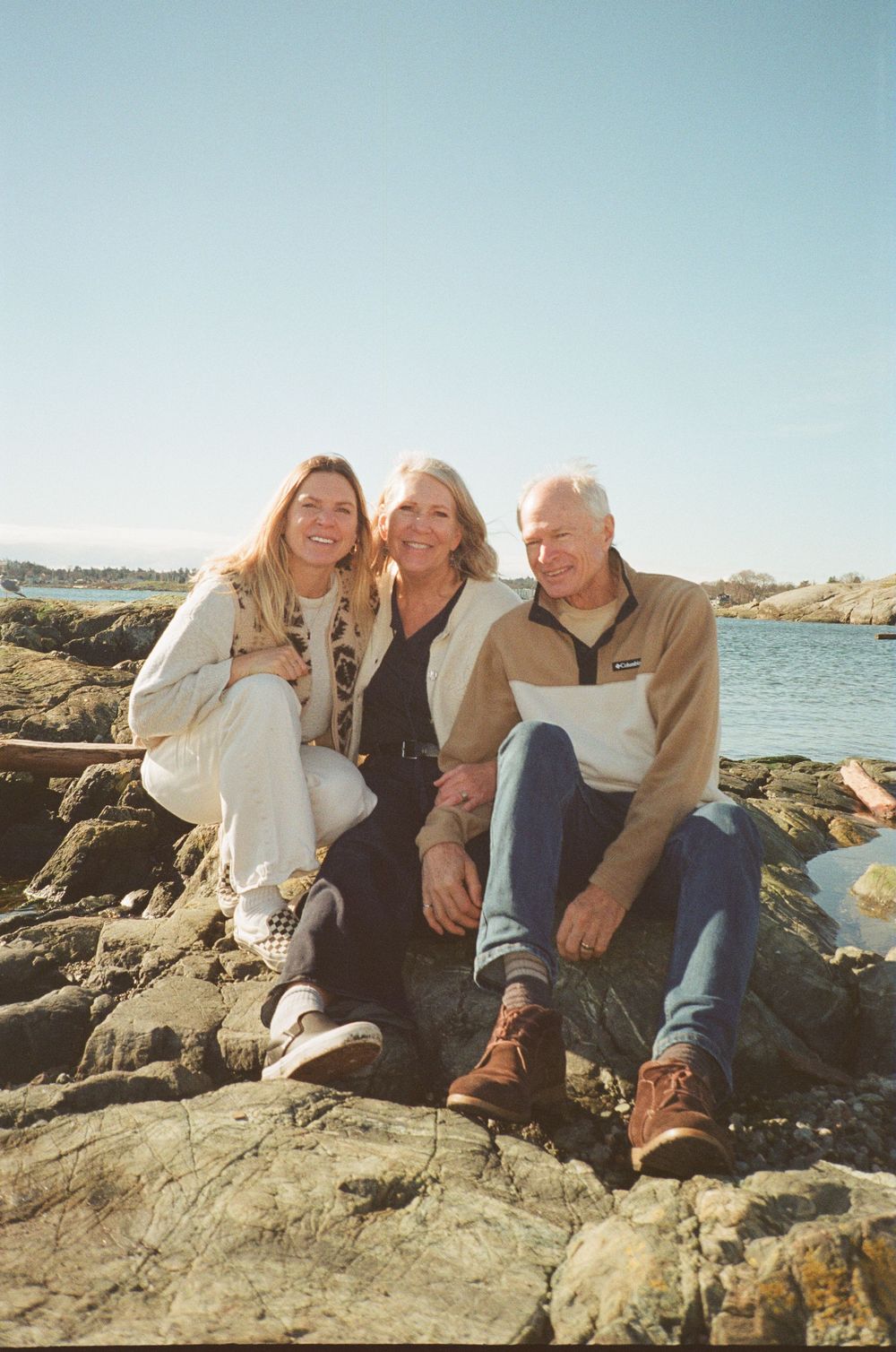 Three adults smiling together while sitting on coastal rocks on a bright sunny day.