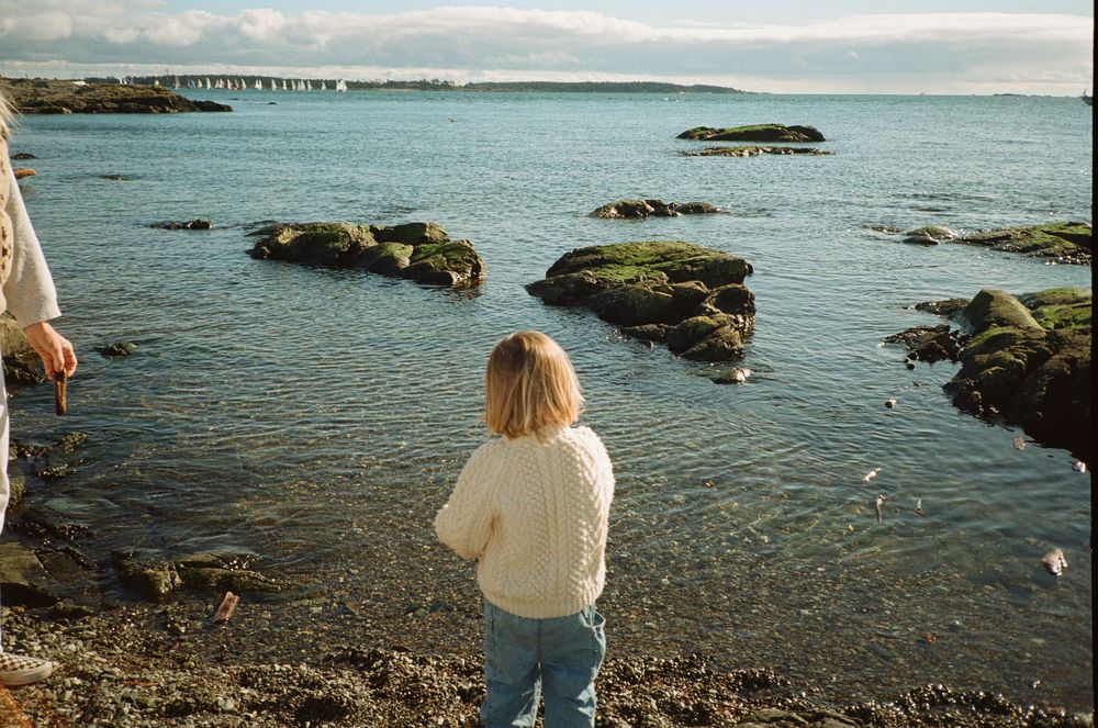 Young blonde child standing at rocky shoreline gazing at calm blue ocean waters.