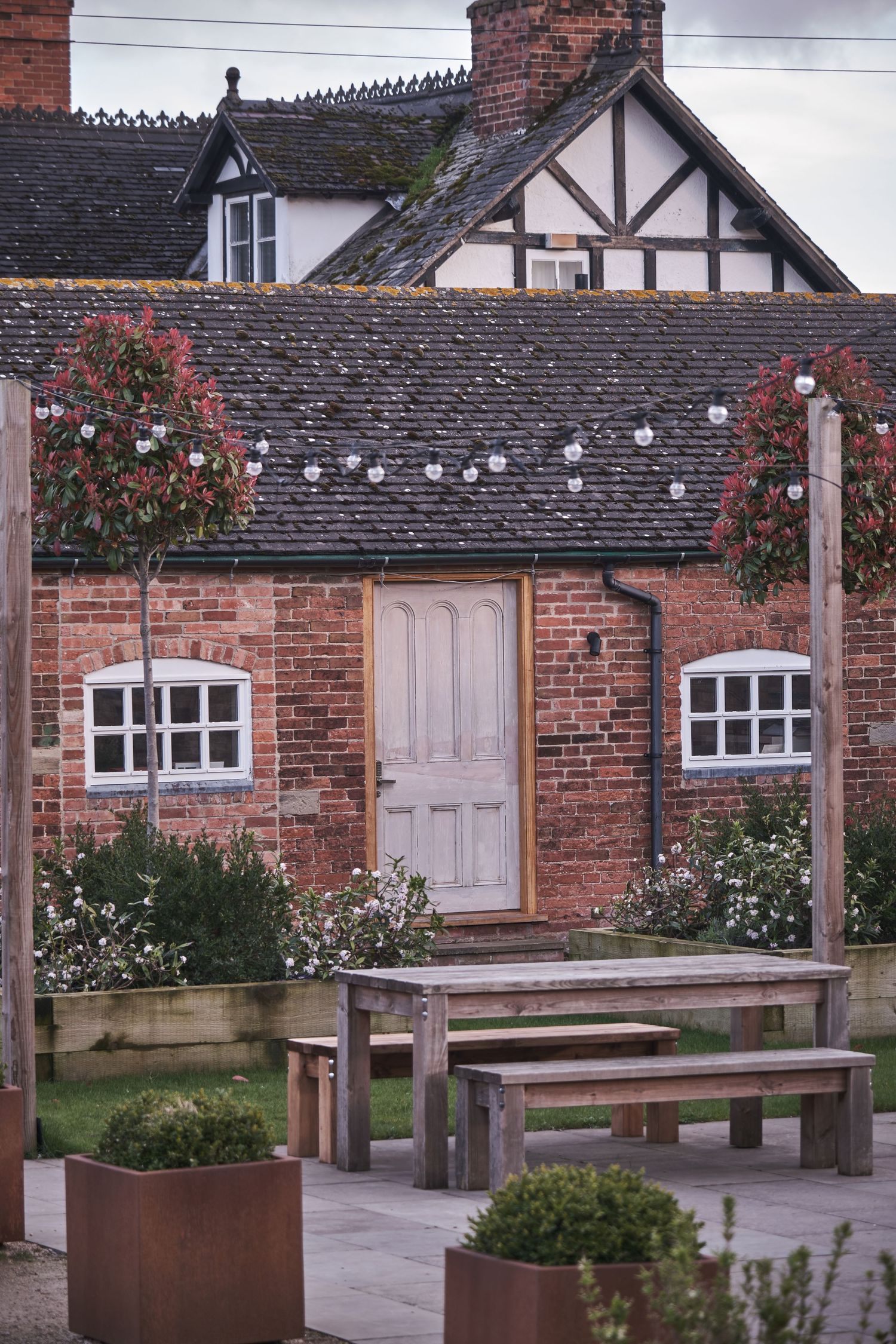 Charming red brick Tudor cottage with string lights, hanging flower baskets, and rustic wooden benches in a paved garden.