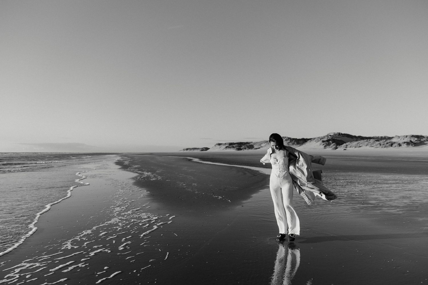 Black and white photo of a woman in white dress dancing freely on a serene beach with gentle waves and sand dunes.