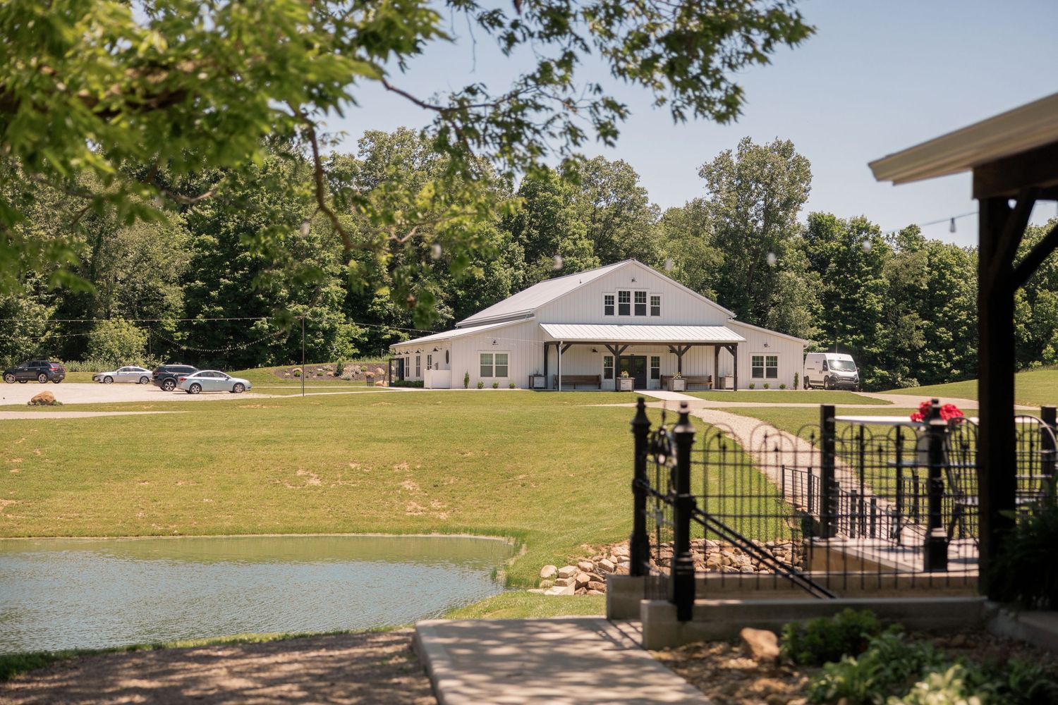 Scenic countryside venue with white barn building, lush green lawn, pond, and black iron fence surrounded by trees. williams farm pataskala