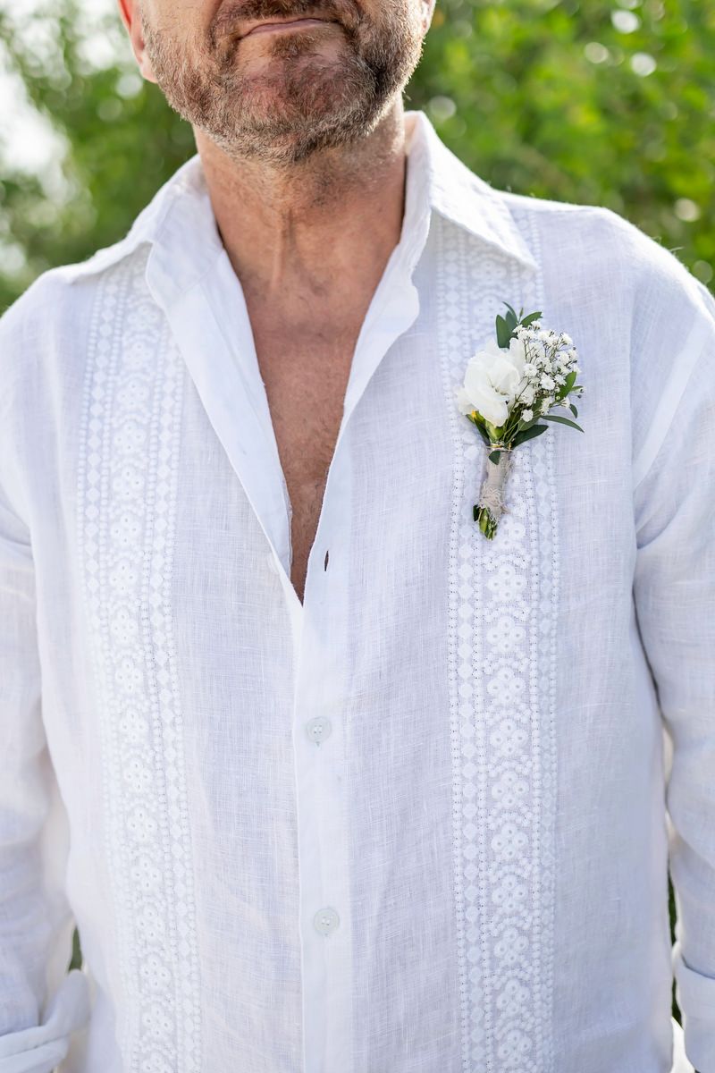 Groom in white linen guayabera shirt with floral boutonniere at outdoor wedding ceremony.