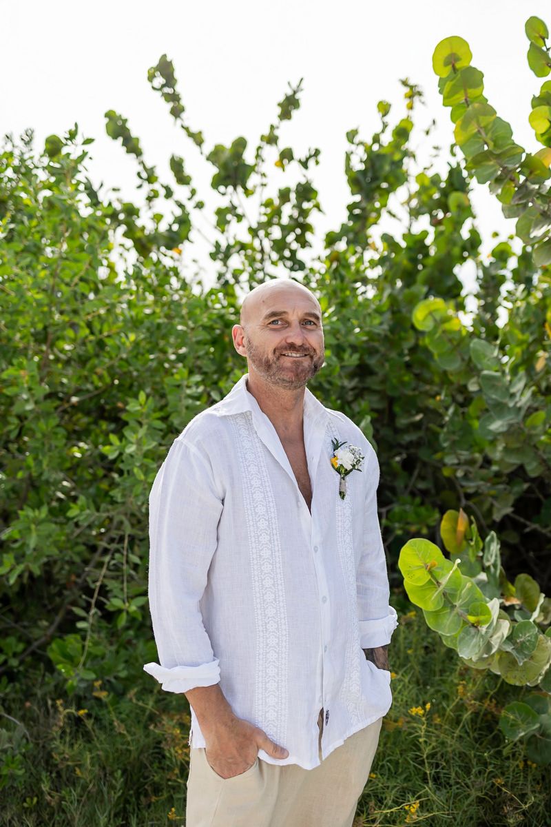 Bald man with beard wearing white linen shirt and boutonniere smiles outdoors among lush green tropical foliage.