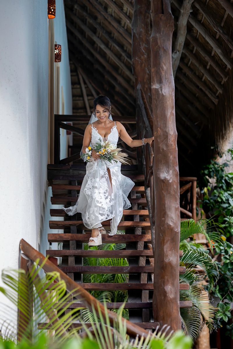 Bride in white lace gown holding bouquet descends wooden stairs at tropical venue with lush greenery.