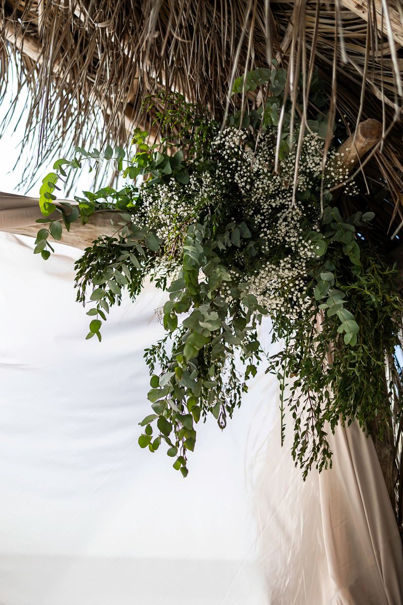 Lush green foliage arrangement hanging from a thatched roof with white draping at an outdoor wedding venue.