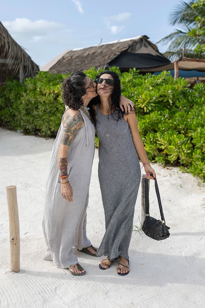 Two women in gray maxi dresses share a kiss on a tropical beach with white sand and lush greenery.