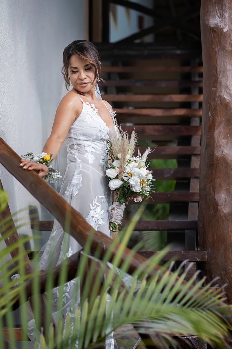 Bride in white lace gown holding floral bouquet on wooden staircase surrounded by tropical plants.
