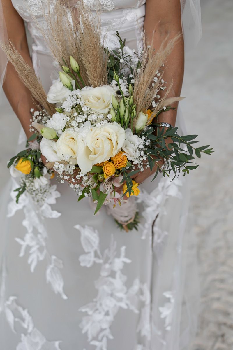 Bride in lace wedding dress holding boho bouquet with white roses, yellow flowers, baby's breath, and pampas grass.