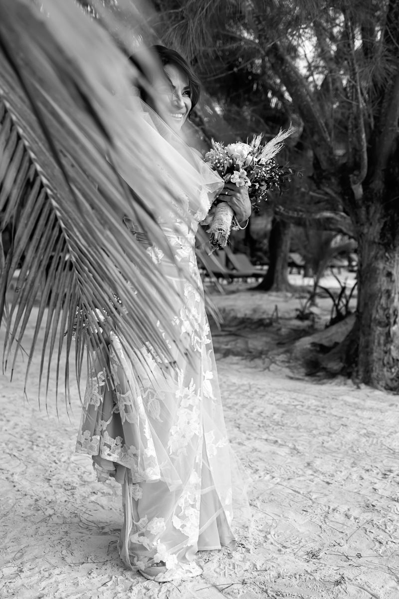 Bride in lace wedding dress holding bouquet under palm frond on sandy beach, black and white photo.