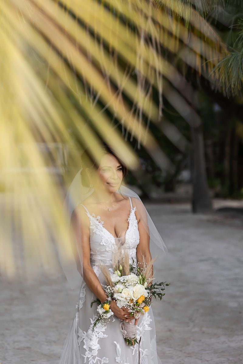 Bride in lace wedding dress holding bouquet of white and yellow flowers, standing beneath palm fronds on sandy beach.