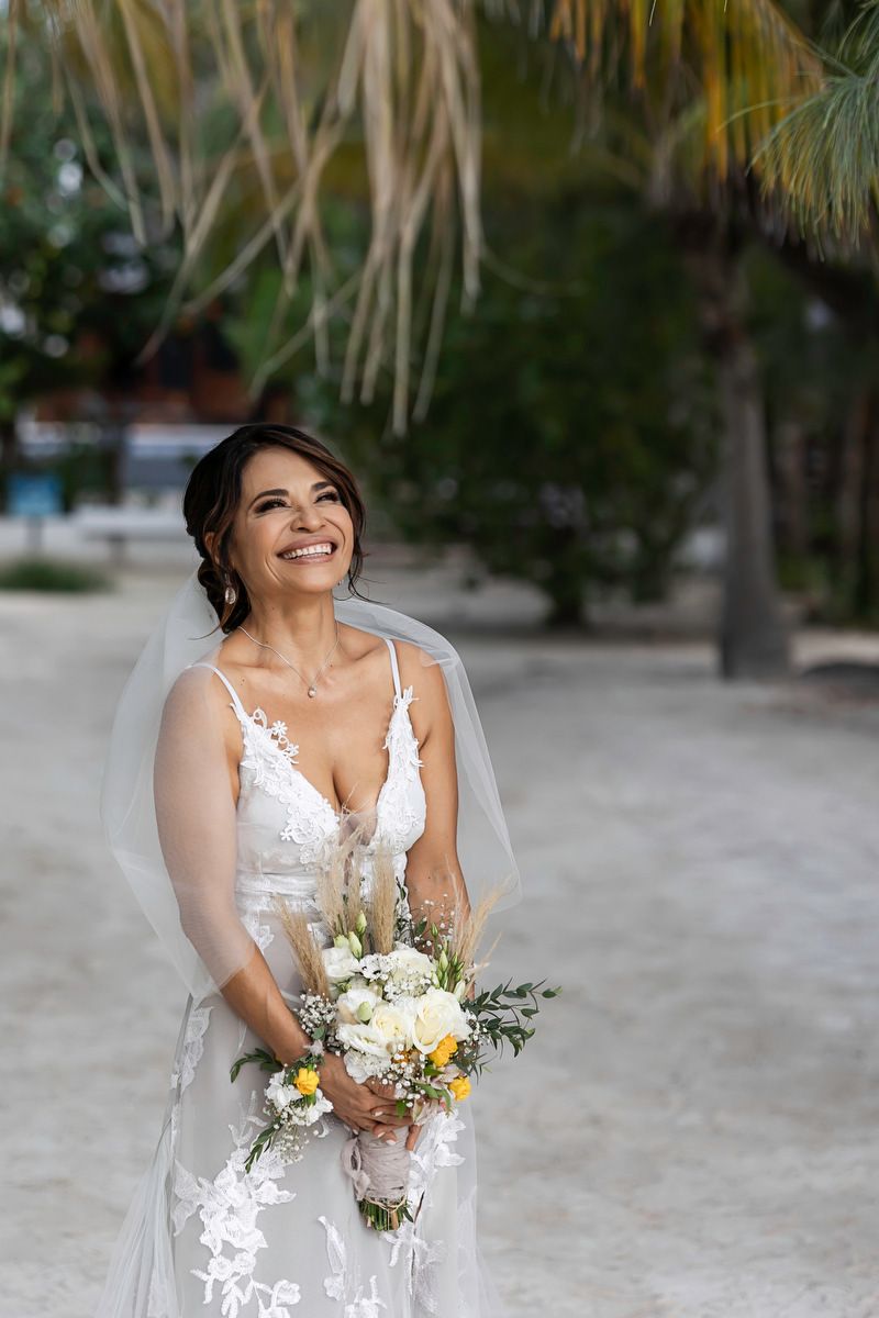 Smiling bride in white lace gown and veil holds bouquet of white and yellow flowers on sandy beach.