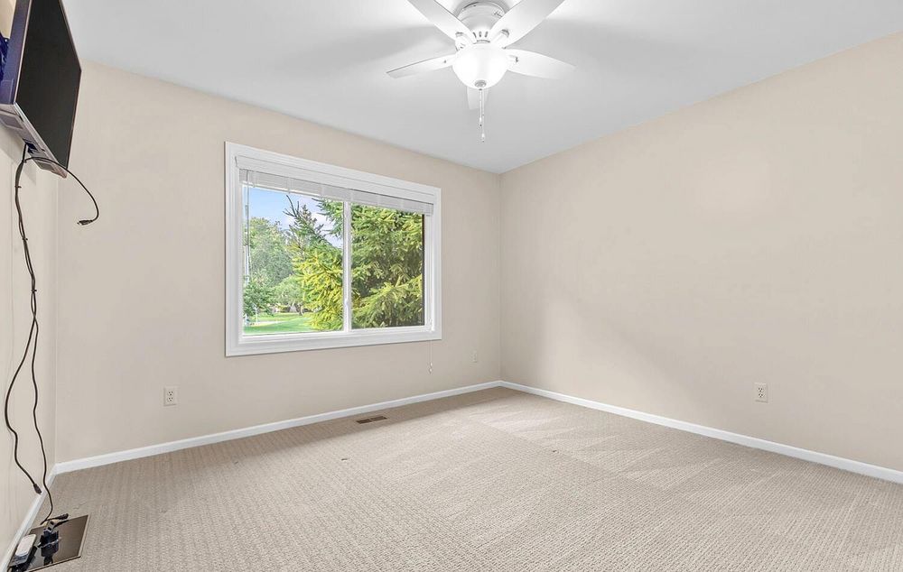 Bright carpeted bedroom with ceiling fan, large window overlooking green trees, and neutral walls.
