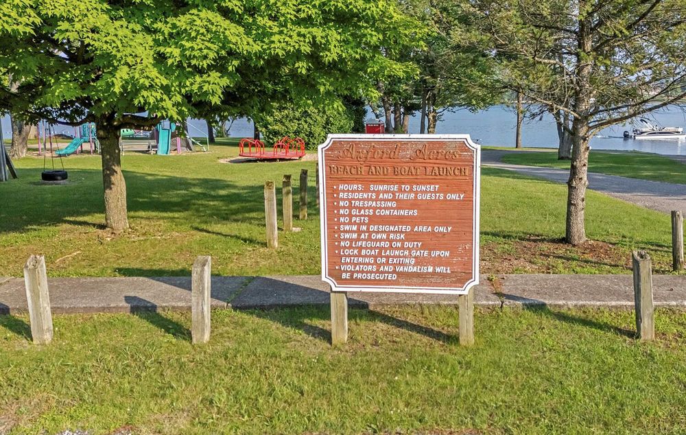 Lakeside park sign with rules posted among mature trees and green grass near the waterfront.