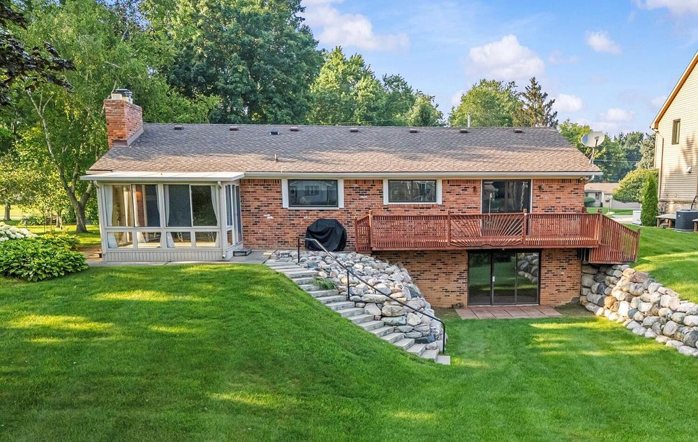 Brick ranch home with wood deck, stone retaining wall, large lawn, and mature trees.