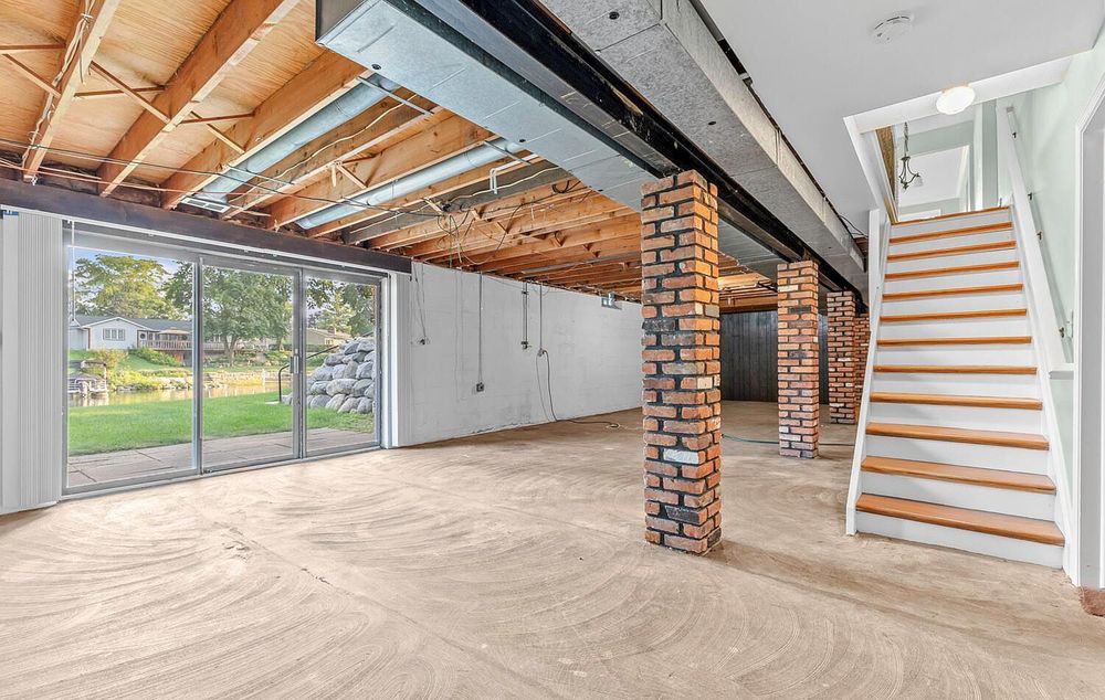 Unfinished basement with brick columns, sliding glass doors, staircase, and open ceiling joists.