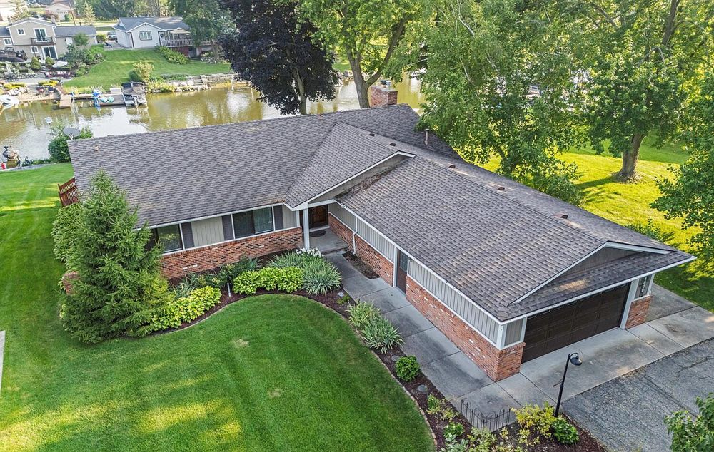 Aerial view of brick ranch home with attached garage, lush lawn, and waterfront setting.