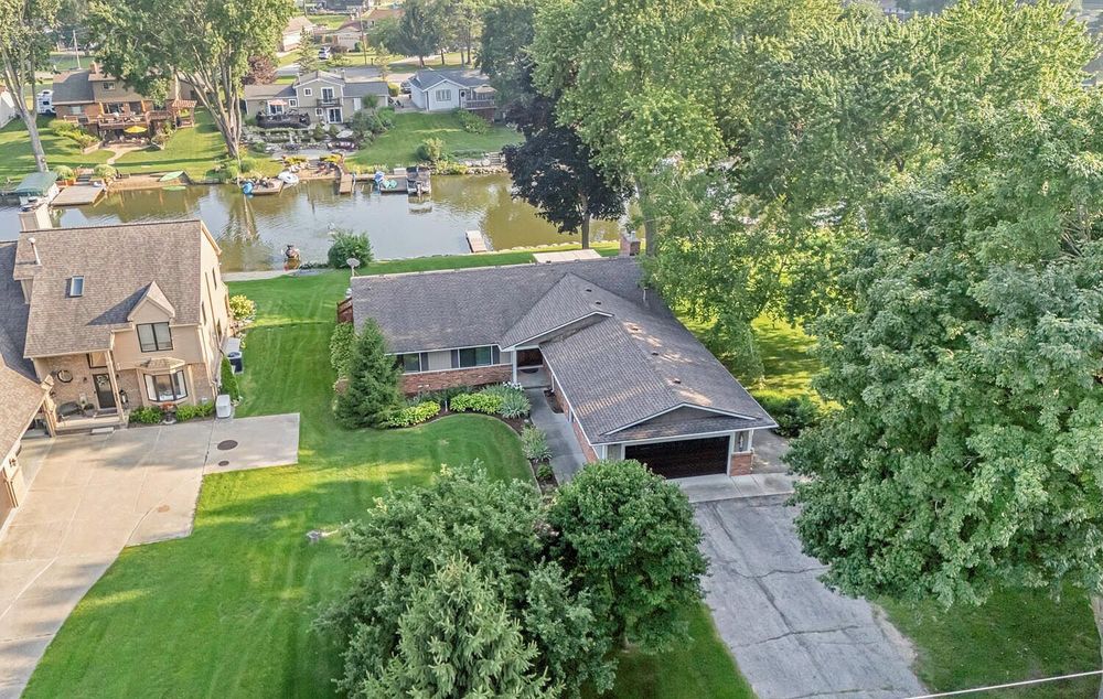 Aerial view of a ranch home with large yard, mature trees, and a scenic pond in the neighborhood.