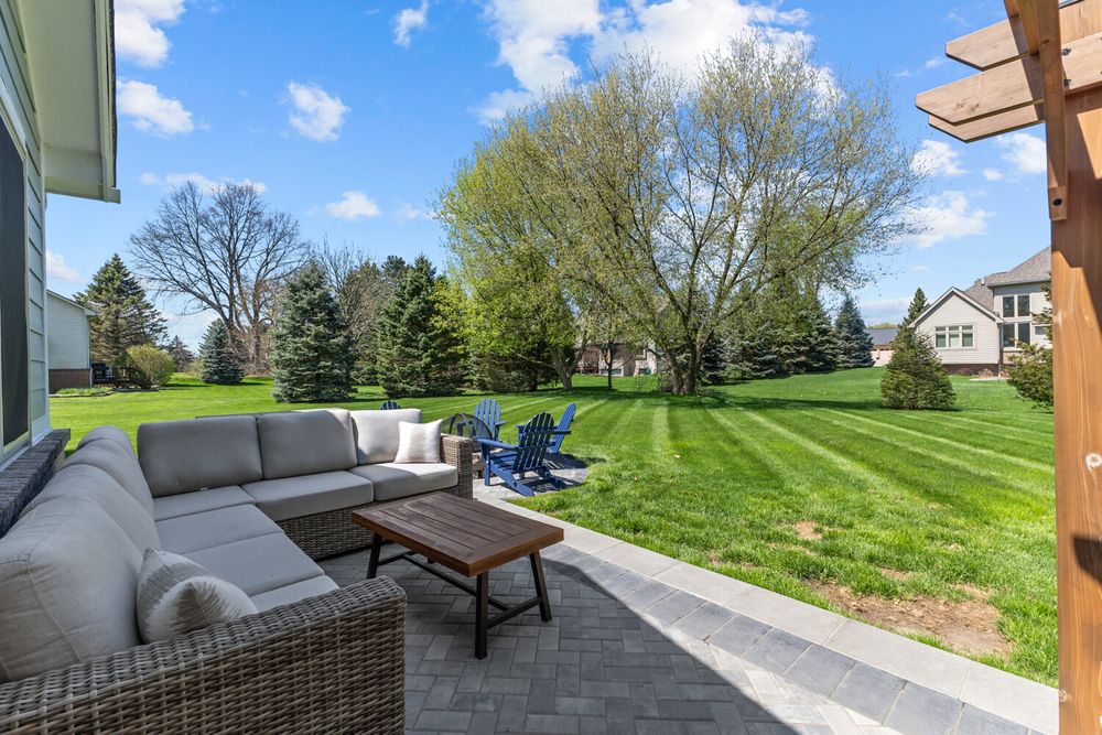 Spacious backyard patio with wicker sectional sofa, Adirondack chairs, and lush green lawn under blue sky.