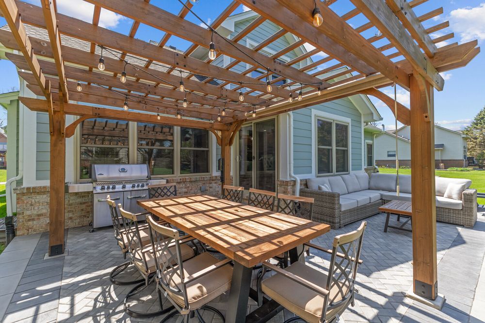 Outdoor pergola patio with dining table, chairs, built-in grill, and wicker seating area on paver surface.