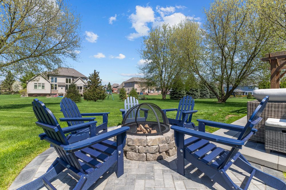 Outdoor patio with stone fire pit surrounded by navy blue Adirondack chairs on a sunny day with lush green lawn.