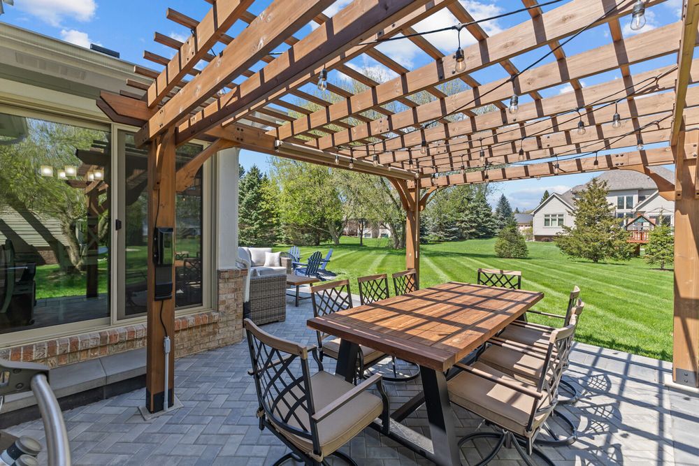 Outdoor pergola patio with string lights, tile-top dining table, and chairs overlooking a lush green backyard.