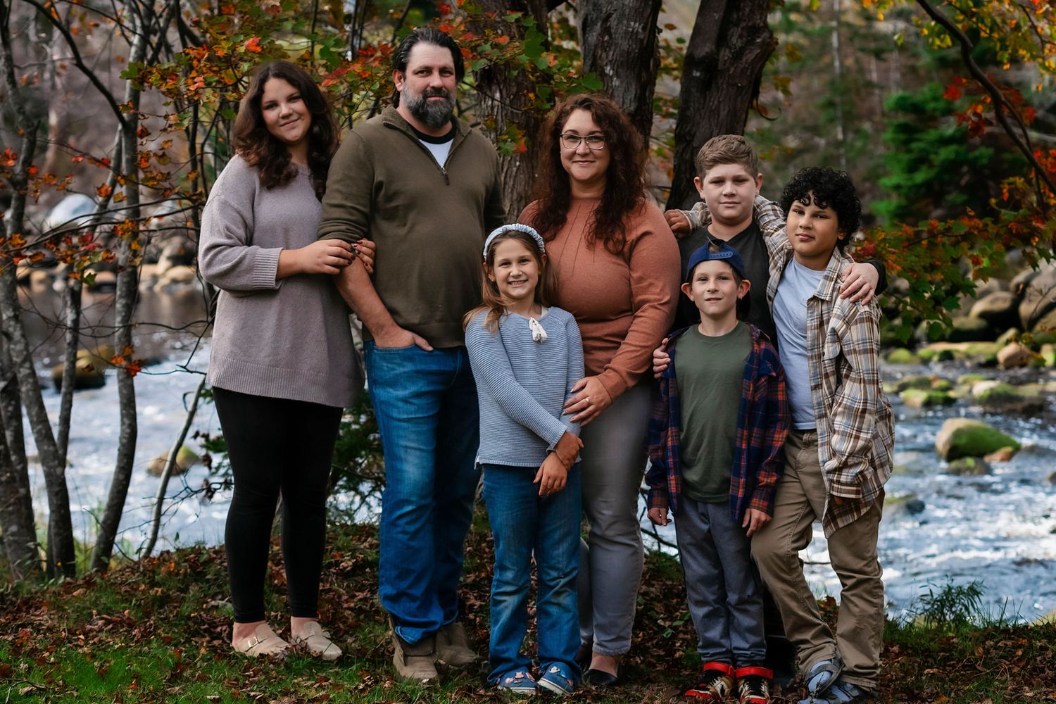 on the Annapolis River a Family of seven posing together outdoors near a rocky stream surrounded by autumn foliage.