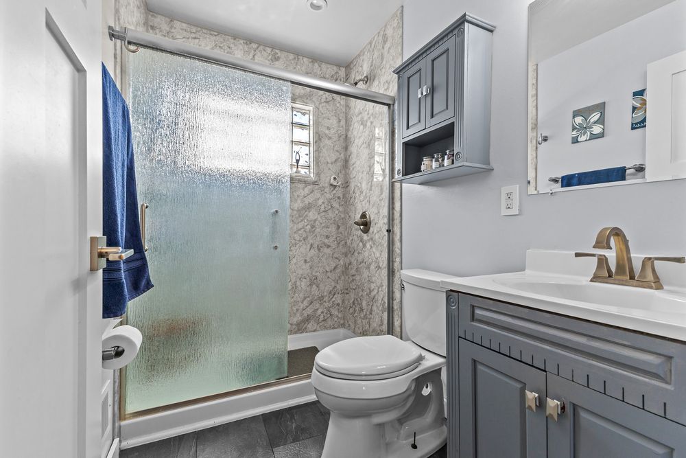 Updated bathroom with gray vanity, gold fixtures, glass shower enclosure, and tiled flooring.