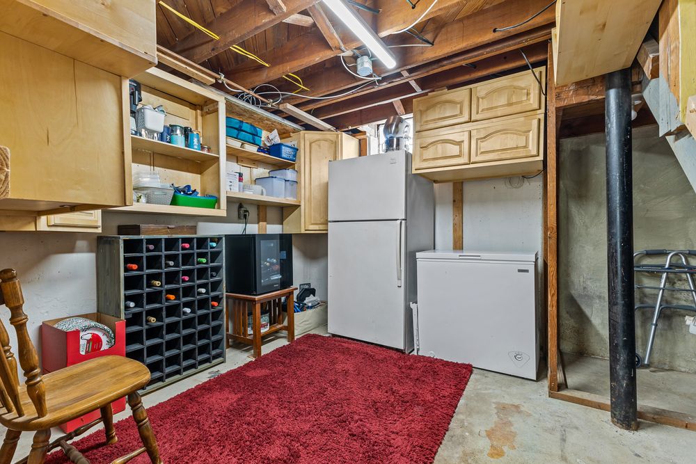 Unfinished basement storage area with wooden shelving, wine rack, refrigerator, and red area rug.