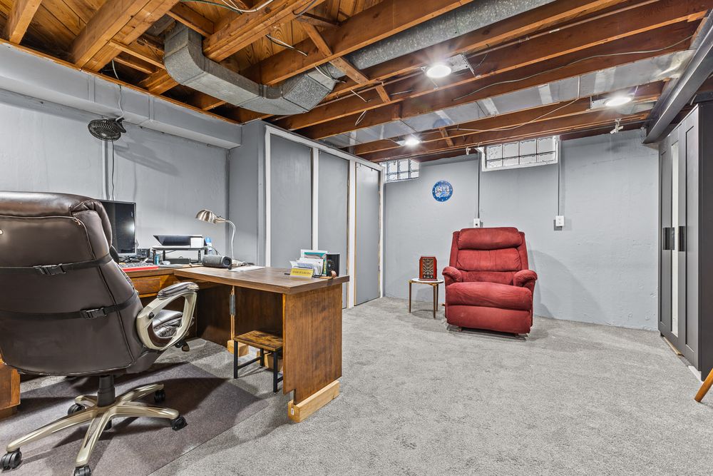 Partially finished basement home office with exposed ceiling, gray carpet, desk, and red recliner chair.